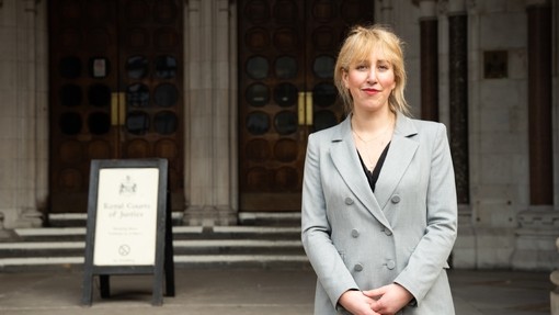 A women dressed in a smart grey blazer stood outside the entrance of a concrete building with two wooden doors.