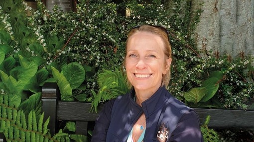 An image of a blonde women wearing a dark blue fleece with The Donkey Sanctuary logo, sat on a bench in front of flowers.