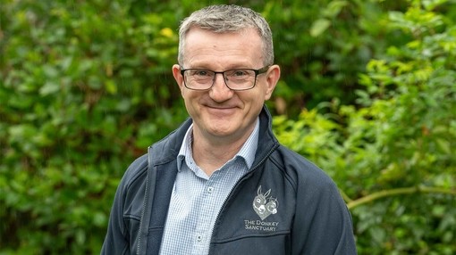 An image of a man wearing glasses and a shirt with a dark blue fleece featuring The Donkey Sanctuary logo smiling toward camera.