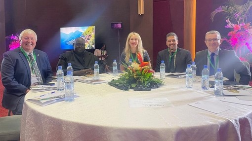 An image of five people sat at a large circular table looking towards camera and smiling.
