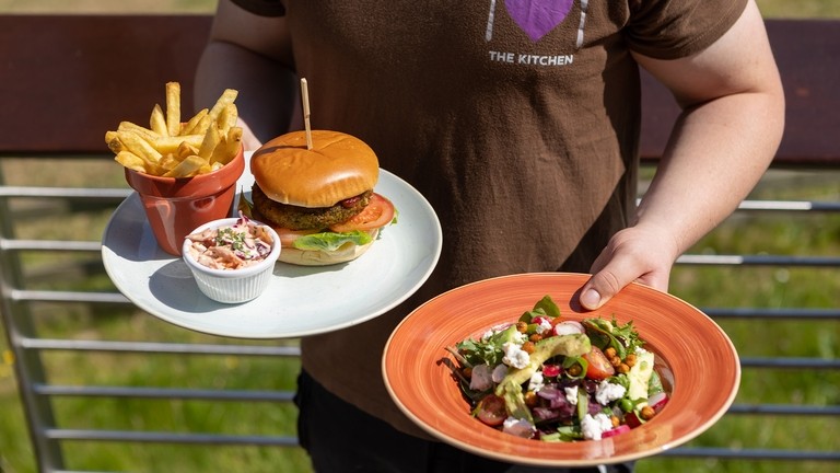 An image of a person wearing a brown t-shirt holding a white plate with a burger and chips in their left hand and an orange plate with a salad in their other hand.