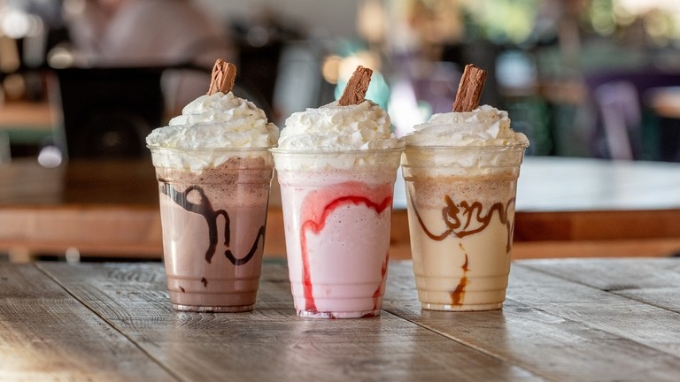 A close up of a brown milkshakes, a pink milkshake and a beige milkshake in plastics cups with whipped cream and a flake in the top
