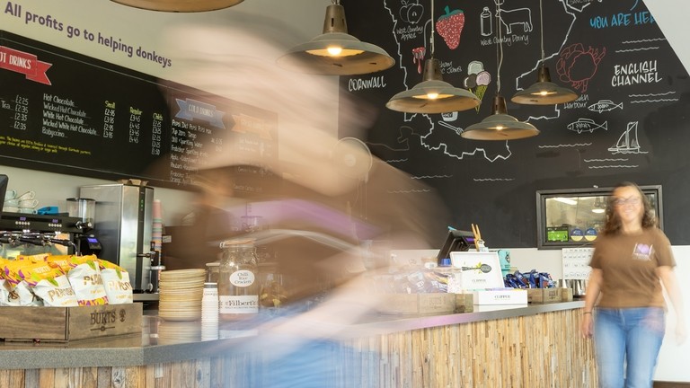 An image of a two people walking in front of a wooden counter with tills on it and a chalkboard with menu items behind.