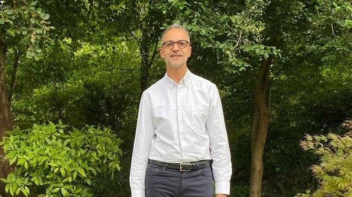 An image of a man wearing glasses and a white shirt, smiling toward camera in front of an area of trees.