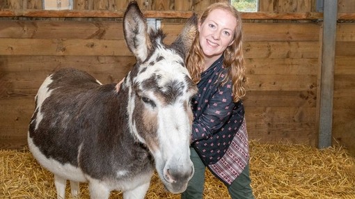 An image of a women inside a wooden shelter stroking a white and grey donkey while smiling at the camera.