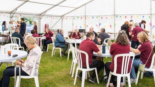 An image of groups of people wearing burgundy tops sat at circular tables under a white marquee.