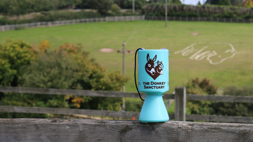 An image of a blue money collection tin resting on top of a fence with a large field and trees in the background.